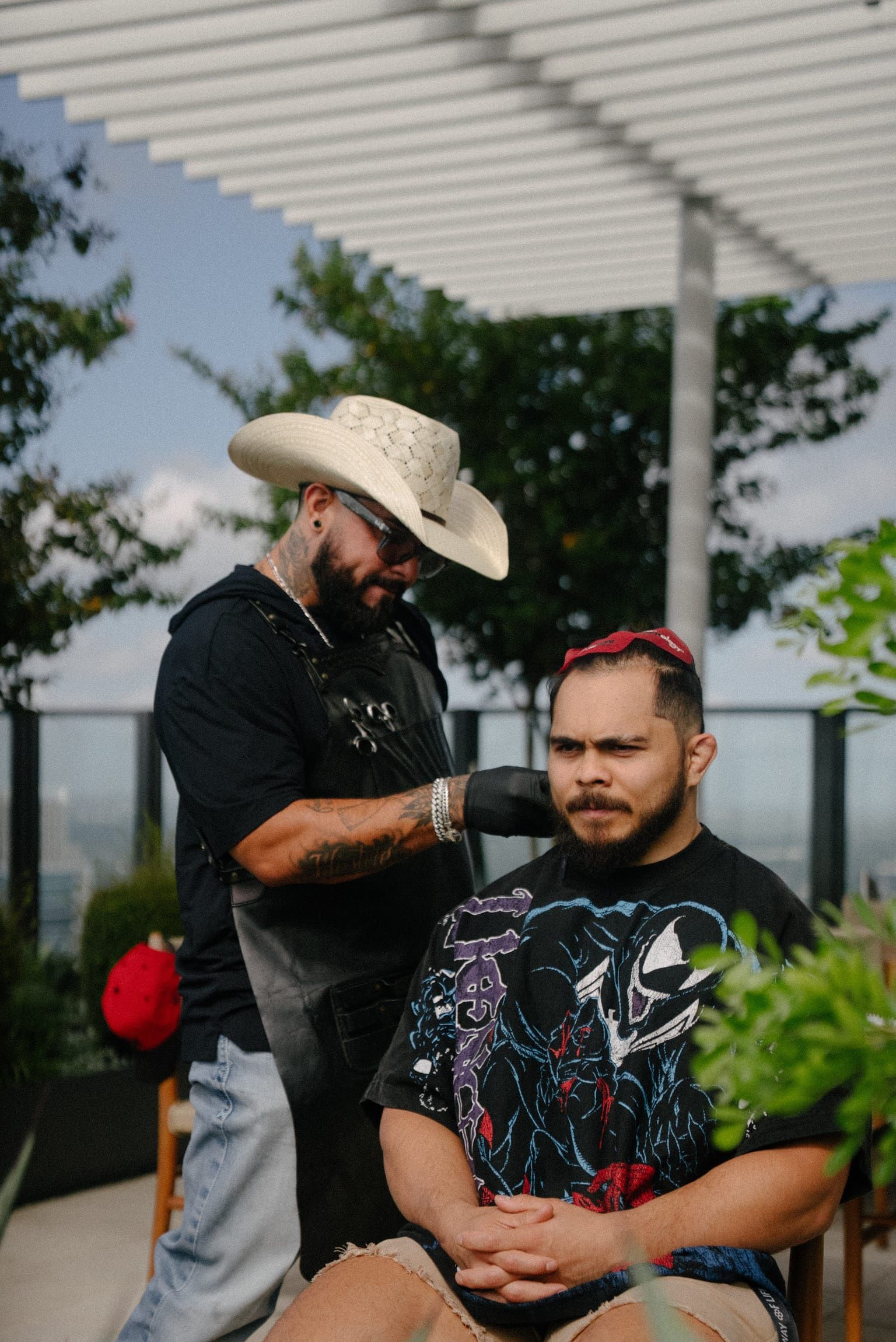 Roy in a straw cowboy hat, shaping a client's beard on a rooftop terrace